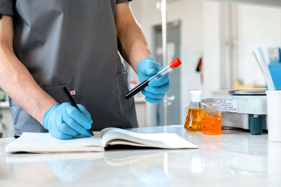 Scientist wearing gloves records lab observations, holding test tube and writing in a notebook.